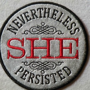 May include: A silver and black circular embroidered patch with the text "NEVERTHELESS SHE PERSISTED" in black and red lettering.