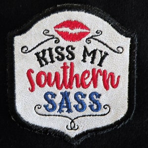May include: White embroidered patch with black, red, and blue lettering. The patch says "Kiss My Southern Sass" with a red lip design above the text.