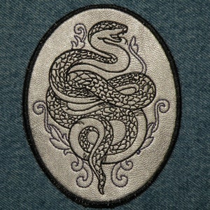 May include: A silver oval-shaped patch with a black embroidered snake design. The snake is coiled and has a detailed scale pattern. The patch has a black border.