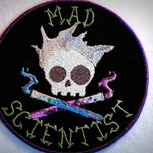 May include: A black and purple embroidered patch with a white skull and crossbones. The skull has a white mohawk and the crossbones are crossed over a blue and purple potion. The text "MAD SCIENTIST" is written around the skull and crossbones.