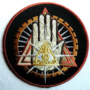 May include: A round embroidered patch with a black background. The patch features a white hand with an eye inside a triangle, surrounded by a gold and brown gear-like design.