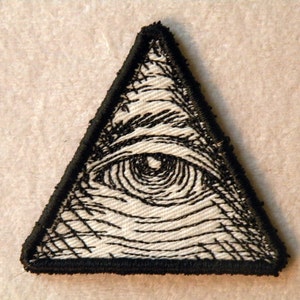 May include: A black and white embroidered patch of an eye inside a triangle. The eye is detailed with a black pupil and white iris. The triangle has a black border.