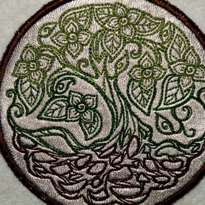 May include: A circular embroidered patch with a green tree design. The tree has a thick trunk and many branches with leaves. The roots of the tree are visible at the bottom of the patch.