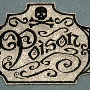 May include: A silver-colored patch with black embroidered details. The patch features the word "Poison" in a gothic font, a skull and crossbones, and decorative swirls. The patch has a textured appearance and a black border.
