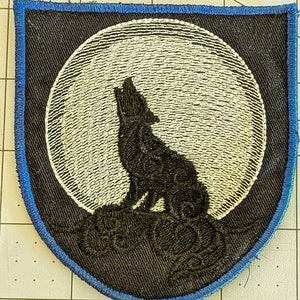 Könnte beinhalten: Ein schwarzer gestickter Aufnäher mit einem Wolf, der zum Mond heult. Der Mond ist weiß und der Wolf ist vor ihm silhouettiert. Der Aufnäher ist von einem blauen Rand umgeben.