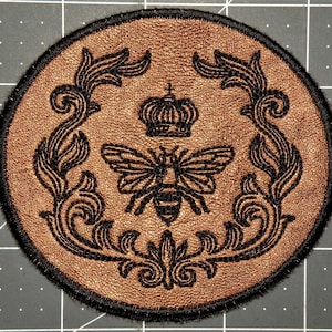 Könnte beinhalten: Runder, brauner Aufnäher mit schwarzem Stickdesign. Das Design zeigt eine Biene, eine Krone und verzierte florale Elemente. Der Aufnäher hat einen schwarzen Rand und ist für Kleidung oder Accessoires geeignet.