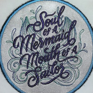 May include: A round, white leather patch with a blue border. The patch is embroidered with the text "Soul of a Mermaid Mouth of a Sailor" in purple thread. The text is surrounded by a decorative design of green and blue thread.
