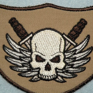 May include: A tan fabric patch with a white skull and crossbones design. The skull has black eyes and a brown nose. The crossbones are brown and have a white wing design on each side.