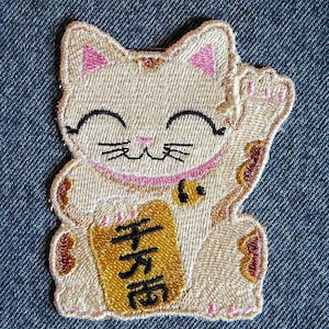 May include: A beige embroidered lucky cat with a raised paw, holding a gold sign with the Japanese character for "fortune".