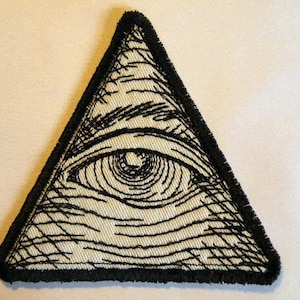 May include: A black and white embroidered patch of an eye inside a triangle. The eye is detailed with a black pupil and white iris. The triangle has a black border.