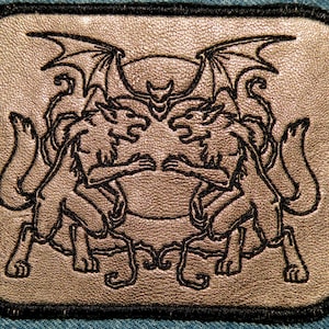 May include: A silver leather patch with a black embroidered design of two mythical creatures with wings and tails, facing each other.