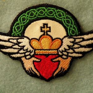 May include: An embroidered patch with a red heart, a gold crown, and white wings. The heart is surrounded by a green Celtic knot design.