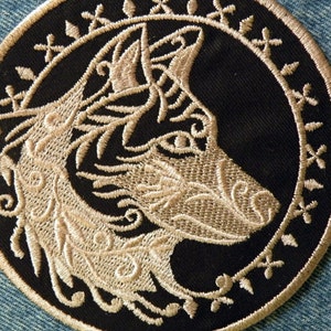 May include: A circular embroidered patch with a black background and a gold embroidered wolf's head with intricate details. The wolf's head is surrounded by a gold embroidered border.