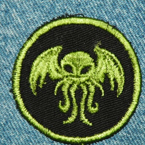 May include: A black and green embroidered patch featuring a stylized depiction of a creature with tentacles and wings. The patch is circular and has a green border.