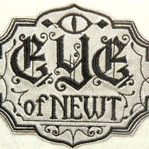 May include: A silver and black embroidered patch with the words "Eye of Newt" in a gothic font. The patch features a stylized eye with a diamond shape above and below it.