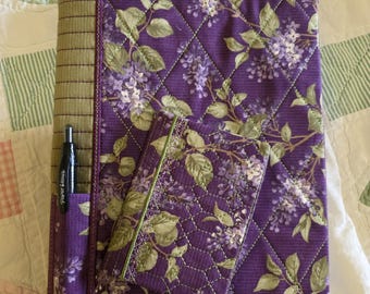 Könnte beinhalten: Ein gesteppter Notizbuchumschlag und ein kleines Notizbuch-Set. Der Umschlag ist tiefviolett mit einem Rautenmuster und einer Stifttasche. Das florale Design zeigt grüne Blätter und weiße und violette Blüten. Das Notizbuch hat die passende Größe.