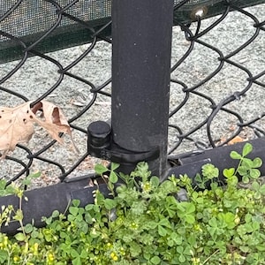 May include: A black metal fence post with a circular black cap and a black mounting bracket attached to a chain-link fence. A single dried brown leaf is caught in the diamond-shaped wire mesh near the base of the post, above green clover.