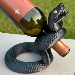 May include: A black and teal snake wine bottle holder. The snake is coiled around the bottle, with its head raised. The wine bottle has a brown label with the word "TALLEROS" on it.