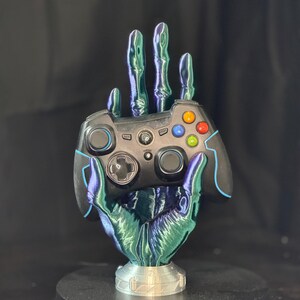 May include: A black video game controller rests in a hand-shaped stand. The stand has a metallic, iridescent finish with shades of green, blue, and purple. The controller has colored buttons and a blue accent. The stand is on a silver base.