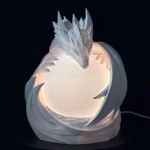 May include: A white dragon sculpture lamp featuring a detailed creature with sharp scales and wings curled around a central, glowing spherical light source. The matte white surface contrasts with the warm, soft illumination emanating from the center.