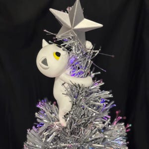 May include: A white cat figurine climbing a silver tinsel Christmas tree, reaching for a white star. The cat has black eyes with yellow pupils. The tree has purple and pink fibre optic lights.