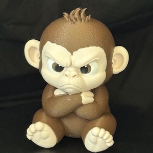 May include: A small, three-dimensional figurine of a brown monkey with a textured surface. The monkey has a cream-colored face, ears, hands, and feet, with a stern, frowning expression and its arms crossed over its chest in a seated position.