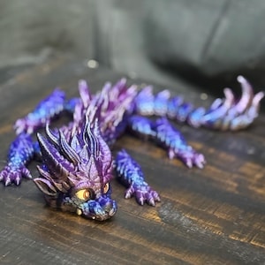 Puede incluir: Figurita de dragón detallada y multicolor en tonos morado, azul y plateado. El dragón tiene una melena puntiaguda, ojos grandes y está posado sobre una superficie de madera. Es un objeto de colección de temática fantástica.