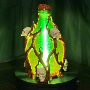 May include: A decorative bottle with a green glowing interior, decorated with brown branches and white skulls. The bottle has a brown cap and sits on a black base. The design has a spooky, Halloween theme.