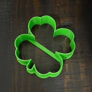 May include: A bright green, three-leaf clover-shaped cookie cutter. The plastic cutter is a solid, vibrant green color and is designed with rounded edges. The clover shape is a classic symbol often associated with St. Patrick's Day.