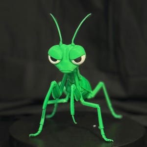 May include: A bright green, articulated praying mantis figurine featuring large, expressive white eyes with black pupils, two long antennae, and segmented legs. The insect model is depicted in a standing position with a focused facial expression.