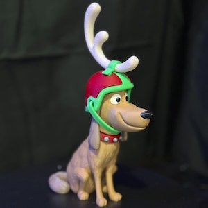 Puede incluir: Figura de un perro con un casco rojo y verde con cuernos blancos. El perro tiene un collar rojo con tachuelas blancas. El perro es de color marrón claro con una nariz negra y una expresión sonriente.