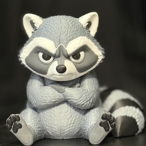 May include: A gray and white raccoon figurine with a textured fur pattern, sitting with its arms crossed over its chest. The raccoon has a stern facial expression, pointed ears, and a striped tail extending to the right side of the frame.