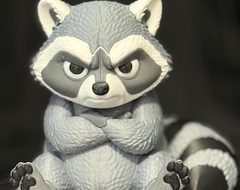 May include: A gray and white raccoon figurine with a textured fur pattern, sitting with its arms crossed over its chest. The raccoon has a stern facial expression, pointed ears, and a striped tail extending to the right side of the frame.