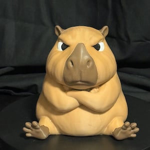 May include: A tan capybara figurine with a dark brown nose and ears, depicted with a stern facial expression and crossed arms. The figure is seated in an upright position with its feet extended forward, featuring a smooth, matte finish throughout.