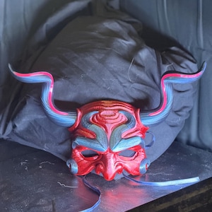 May include: A red, blue, and pink Oni mask with curved horns. The mask features intricate details and a textured surface. Blue ribbons are attached to the sides. The mask is likely for cosplay or costume use.