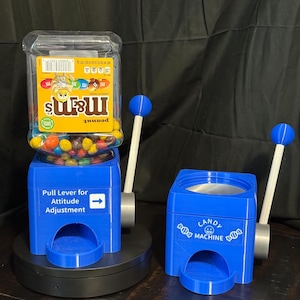 May include: Two blue plastic candy dispensers with white levers and blue spherical knobs. The left dispenser holds an inverted M&M's peanut candy jar with a label reading Pull Lever for Attitude Adjustment. The right dispenser is labeled Candy Machine.