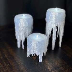 Puede incluir: Tres velas LED blancas cilíndricas con efecto de cera derretida. Las velas, de diferentes alturas, tienen una luz brillante en la parte superior. La cera derretida gotea por los lados, creando una apariencia texturizada única. Dispuestas sobre una superficie de madera oscura.