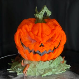 May include: An orange Halloween pumpkin with a carved, sinister face. The pumpkin has a green stem and sits on green leaves. White vapor emanates from the mouth, creating a spooky effect. A festive decoration for Halloween.