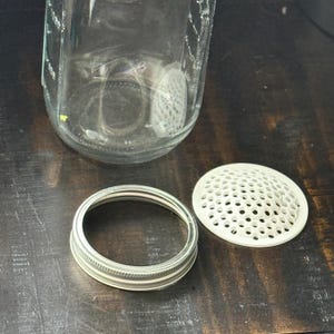 May include: A clear glass jar with a silver metal ring and a white plastic insert with small holes. The jar is on a dark wooden surface. The jar is empty.