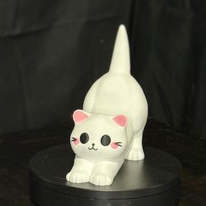 Può includere: Una statuetta di gatto bianco in posa giocosa, con le zampe anteriori distese e la coda sollevata. Il gatto ha orecchie e guance rosa e occhi neri. La statuetta è su una superficie nera.