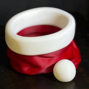 May include: A white, circular object sits atop a red, pouch-like form, with a small white sphere nearby. The white ring has a smooth, rounded shape. The red form has a textured, fabric-like appearance. The objects are on a dark surface.