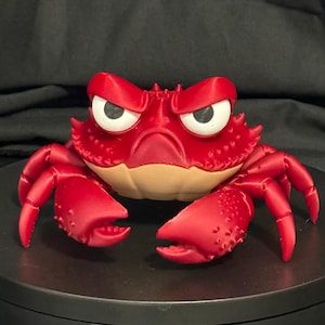 May include: A red crab figurine with a textured shell, large white eyes with black pupils, and a tan-colored underside. The crab has two prominent claws and multiple legs extending from its sides, featuring a stern facial expression.