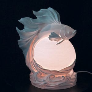 May include: A white sculptural lamp featuring a betta fish with flowing fins positioned above a glowing spherical light source. The base of the lamp is molded into a wave-like pattern, and the entire structure emits a soft, warm light.