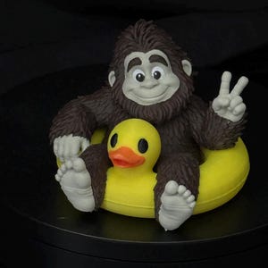 May include: A brown, furry creature with a light gray face, hands, and feet, sitting inside a bright yellow rubber duck floatie. The creature has a smiling expression and is holding up one hand in a peace sign gesture, with black eyes and nose.