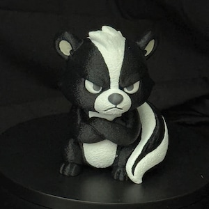 May include: A small, stylized skunk figurine with black and white fur, featuring a prominent white stripe running from its forehead down its back. The skunk has a grumpy facial expression with narrowed eyes, a small nose, and arms crossed.