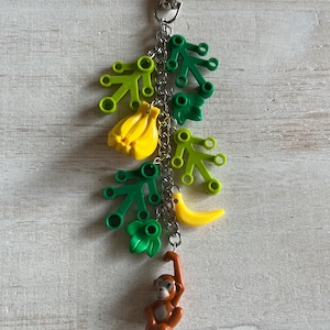 May include: A whimsical keychain featuring a brown monkey, yellow bananas, and green leaves. The charm is attached to a silver-colored clasp and chain, creating a playful accessory.