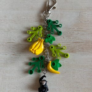 May include: A novelty keychain featuring a silver clasp and chain, adorned with yellow bananas, green leaves, and a small black monkey figurine. The keychain is designed with a playful, tropical theme.