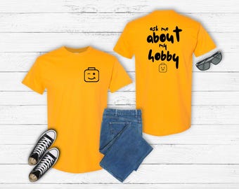 May include: Mustard yellow t-shirts with black graphics and text. One shirt has a winking block figure, the other says "ask me about my hobby" with a block figure. Includes blue jeans and black sneakers.