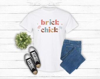 May include: White t-shirt with the text "brick chick" in colorful letters. The shirt is paired with blue jeans and black and white sneakers. A small green plant is also in the image.