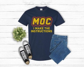 May include: Navy blue t-shirt with the text "MOC I MAKE THE INSTRUCTIONS" in yellow and red. The shirt is paired with blue jeans and black and white sneakers. A small green plant is also in the image.